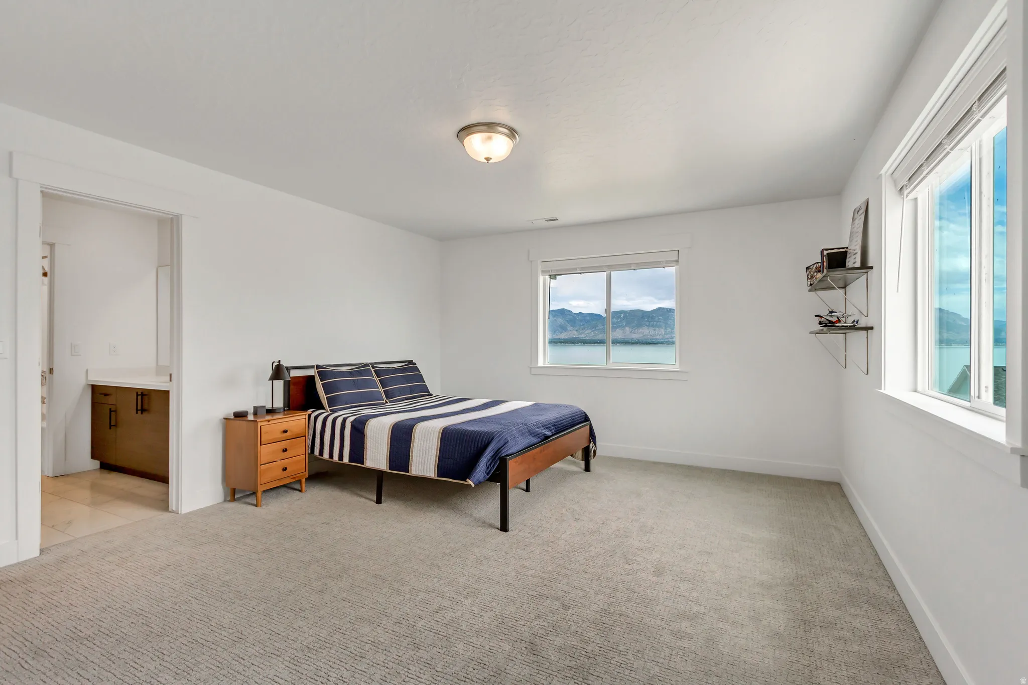 Spacious bedroom has entrance to vanity area then another door to private shower/toilet area, two windows with lake and mountain views, light carpet, and white walls