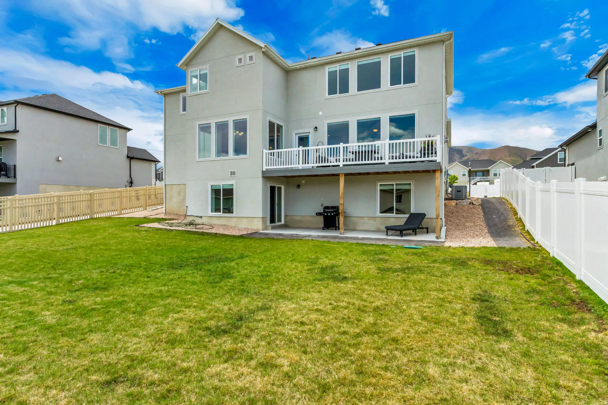 Expansive rear yard featuring a manicured lawn, a full-length balcony with white railing, a ground-level covered patio and vinyl fencing on both sides.