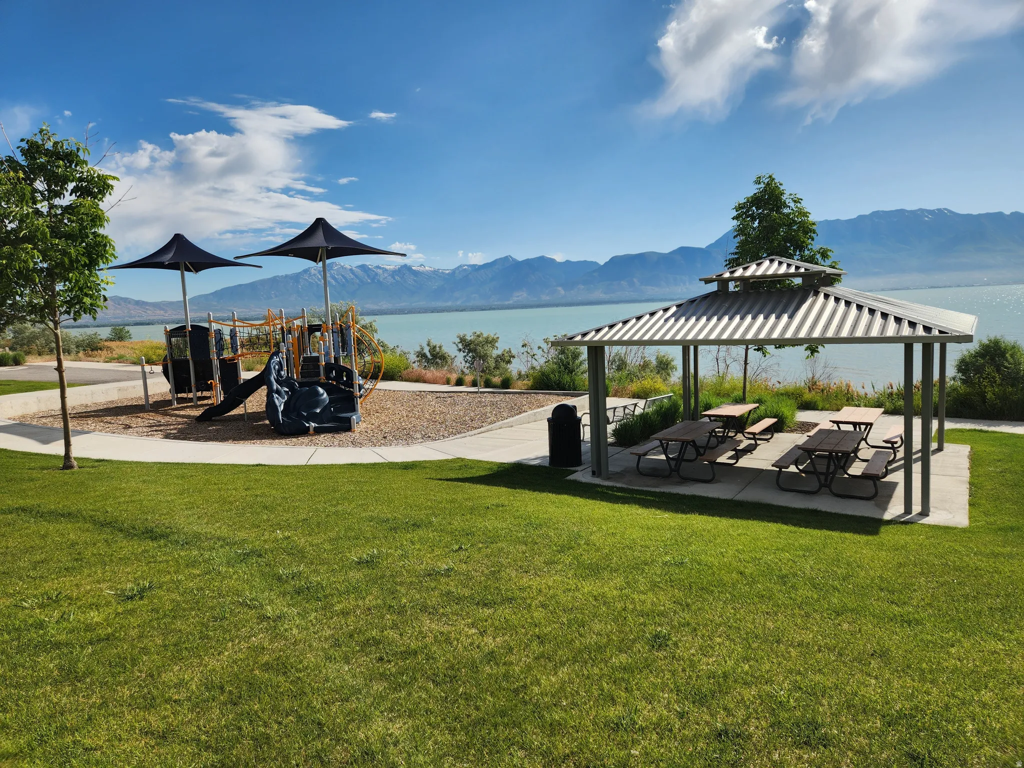 Playground is one block away. Expansive lakeside grassy area featuring a children's play structure with slides and swings, a covered picnic area with multiple tables, and a paved pathway