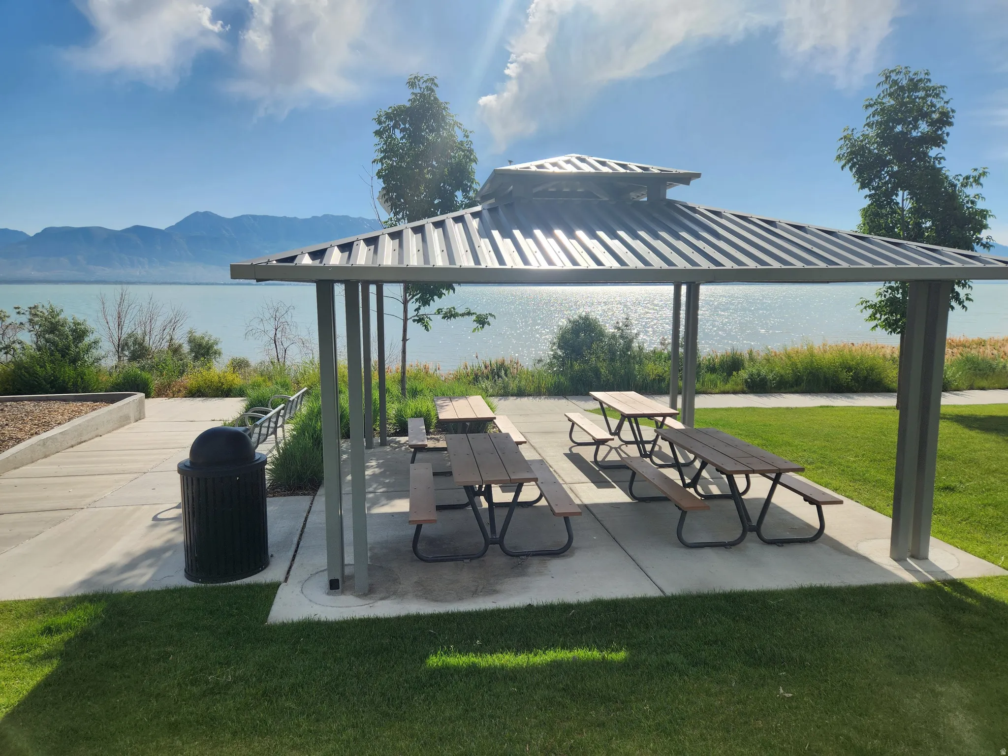 Picnic area is next to the playground. Shaded lakeside picnic area featuring a metal gazebo, multiple picnic tables, and concrete pathways