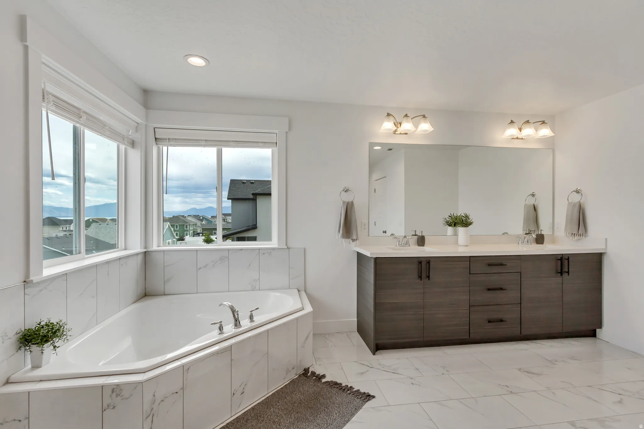 Spacious primary bathroom featuring a corner soaking tub with marble-finish tile surround, large windows, wood-finish double vanity with dual sinks and quartz countertop, and large wall-mounted mirror