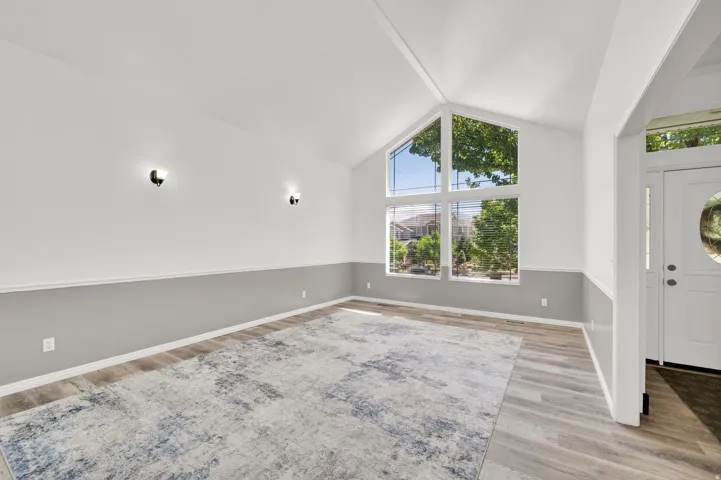 Vaulted ceiling living space featuring a large picture window, wood-finish flooring, two-tone wall treatment, and sconce lighting
