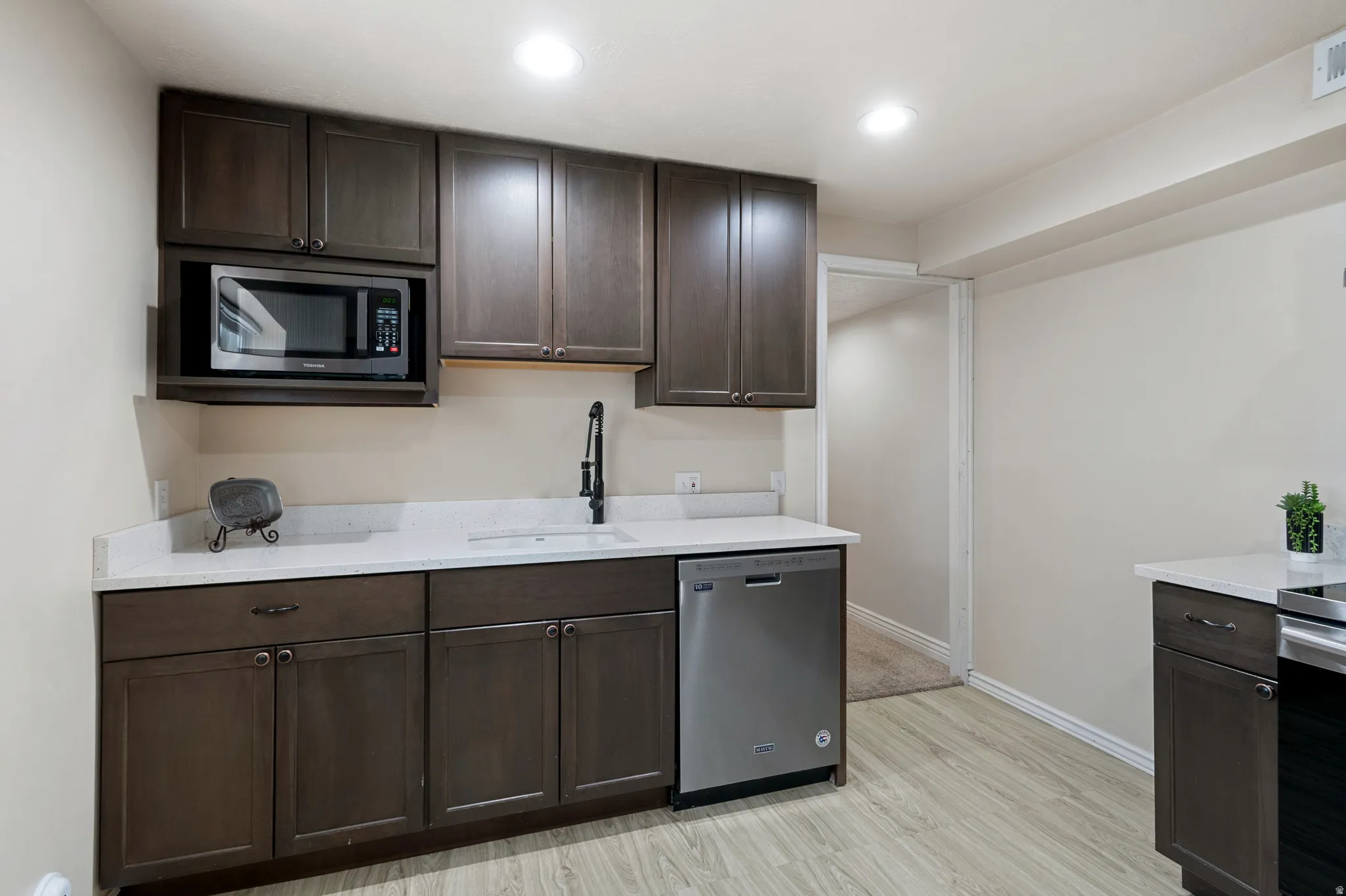 Kitchenette featuring dark wood-finish cabinetry, light-toned countertops, a stainless steel dishwasher, a built-in microwave, and wood-finish flooring