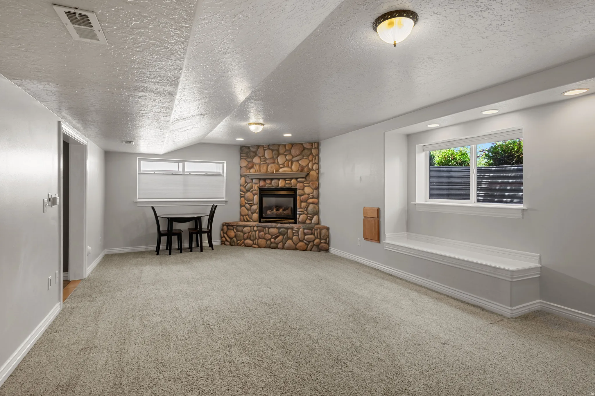 Spacious room featuring light gray carpeting, a stone-clad fireplace, and a built-in window bench