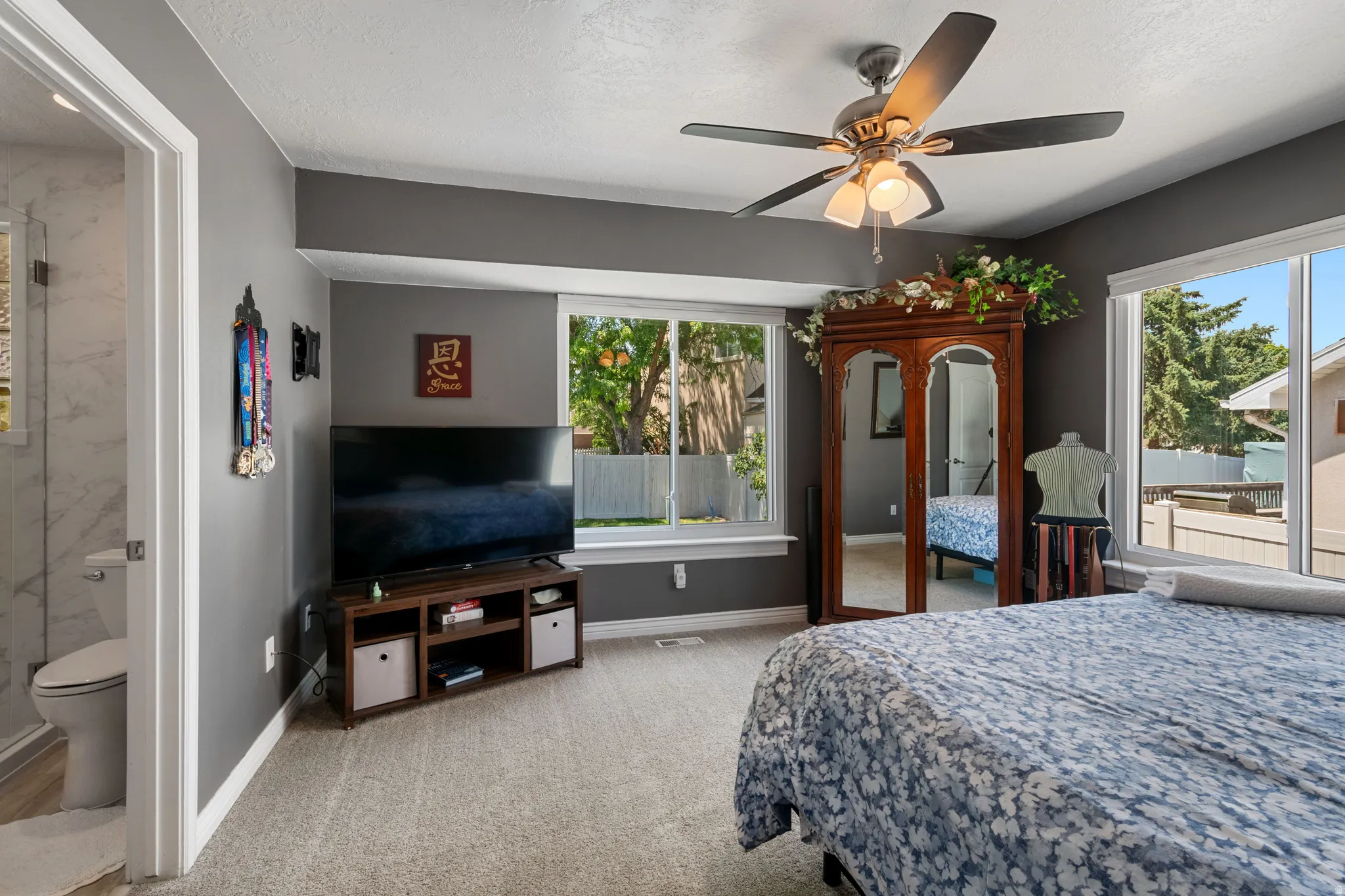 Carpeted room featuring multiple windows, a ceiling fan with integrated lighting, and a doorway opening to a bathroom with a white toilet and patterned wall tile