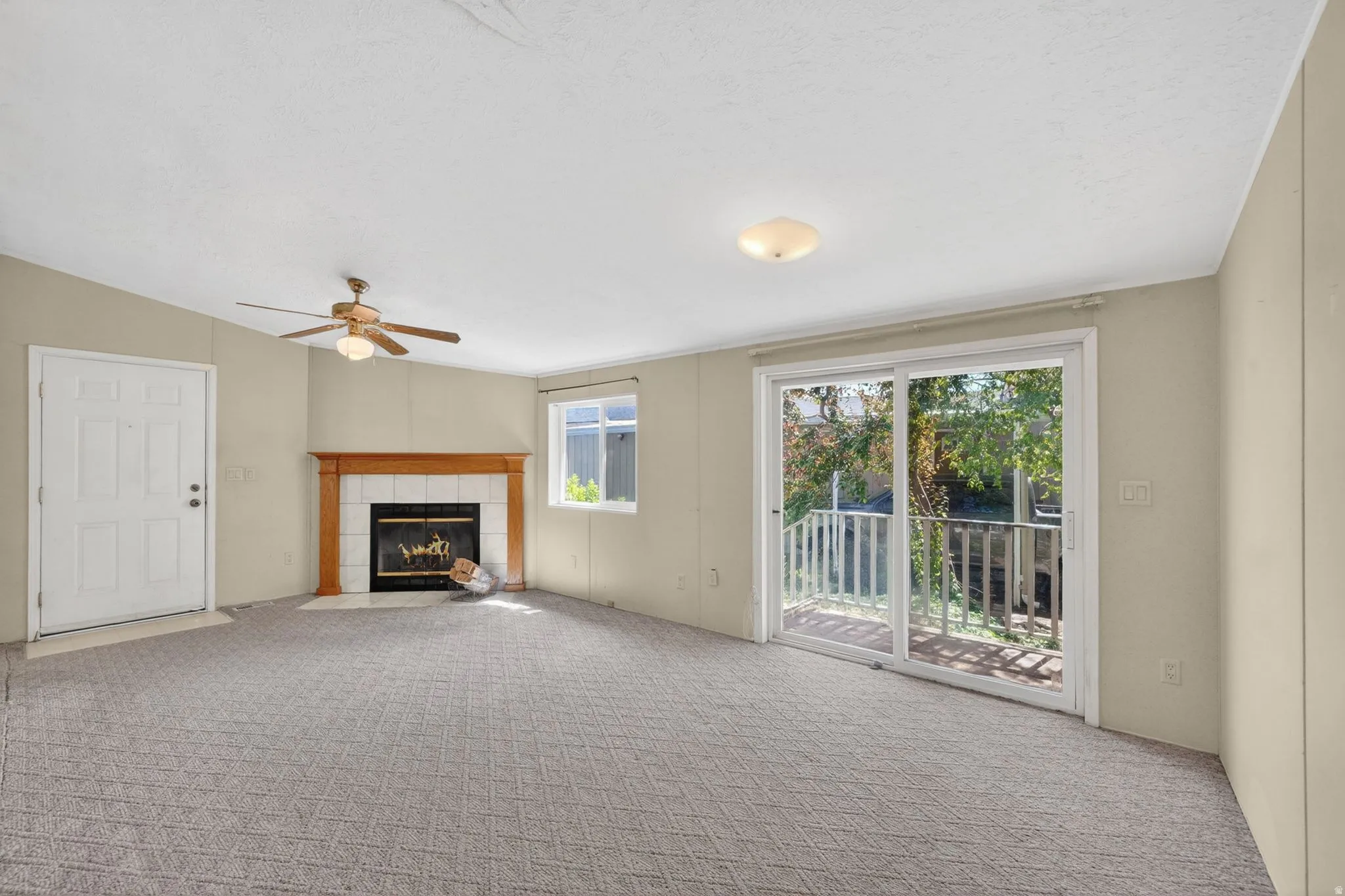Living space featuring a corner fireplace with a wood mantle and tile surround, patterned carpet flooring, a ceiling fan with integrated lighting, a sliding glass door to exterior, and a window providing natural light