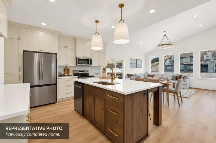 Open-concept living space featuring a central kitchen island with a gold-tone faucet, light wood-finish flooring, vaulted ceilings, and large windows