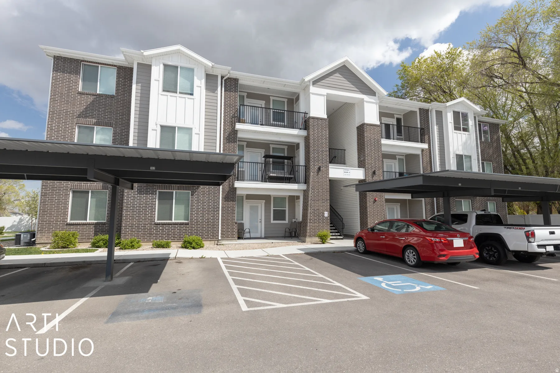 Contemporary multi-story building featuring brick and panel siding, private balconies with black railings, covered parking structures, and a dedicated accessible parking space