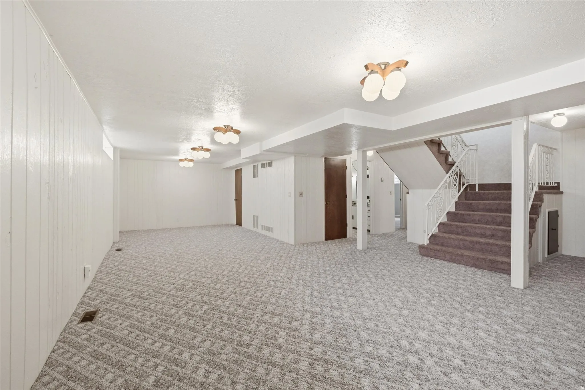 Spacious lower-level area with patterned carpet flooring, a carpeted staircase with a white metal railing, white paneled walls, and multiple ceiling-mounted light fixtures