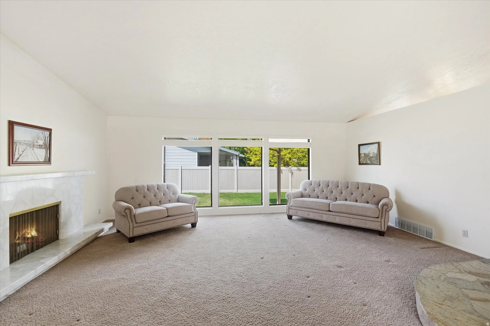 Spacious living area featuring a prominent fireplace with a marble-finish surround, large windows providing natural light, and plush carpet flooring throughout