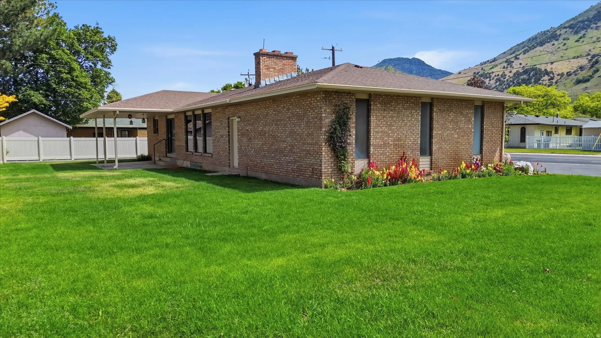 Brick exterior featuring a covered patio, extensive lawn space, mature trees, a garden bed with blooming plants, and a partial mountain backdrop