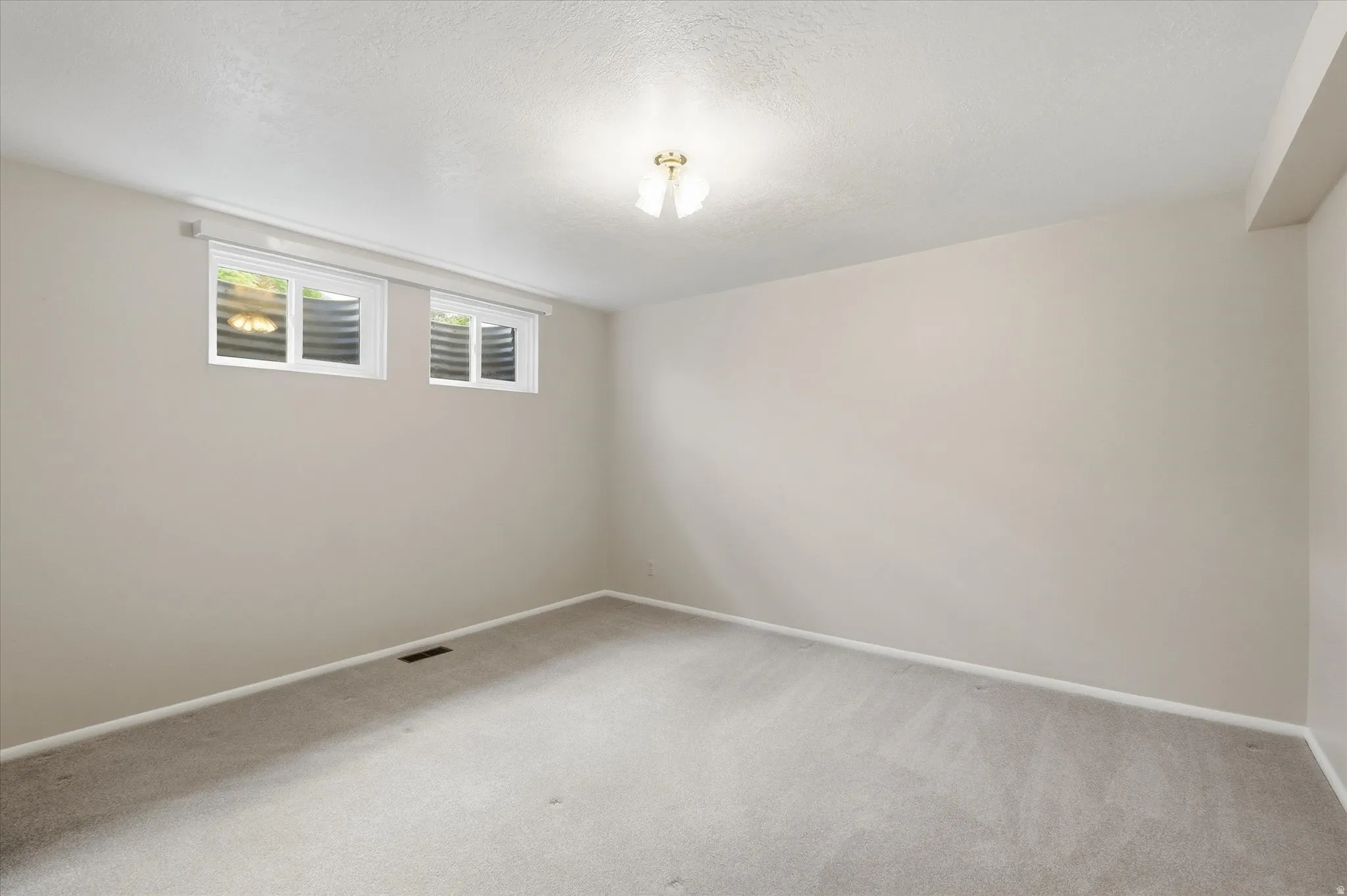 Carpeted room with neutral-toned walls, two basement windows, and a ceiling-mounted light fixture