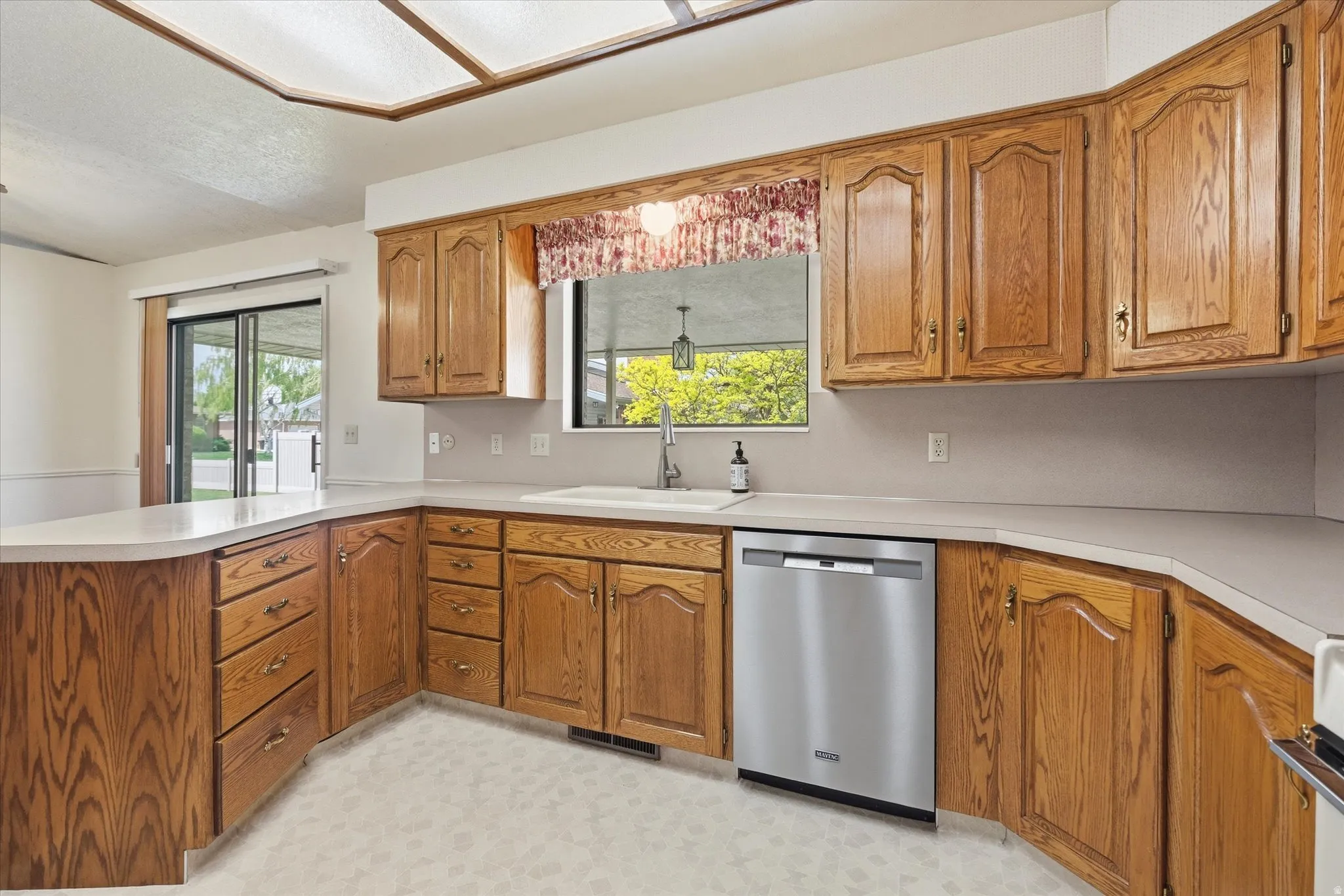 Kitchen featuring wood-finish cabinetry with brass-tone hardware, light-toned countertops, a stainless steel dishwasher, and an undermount sink