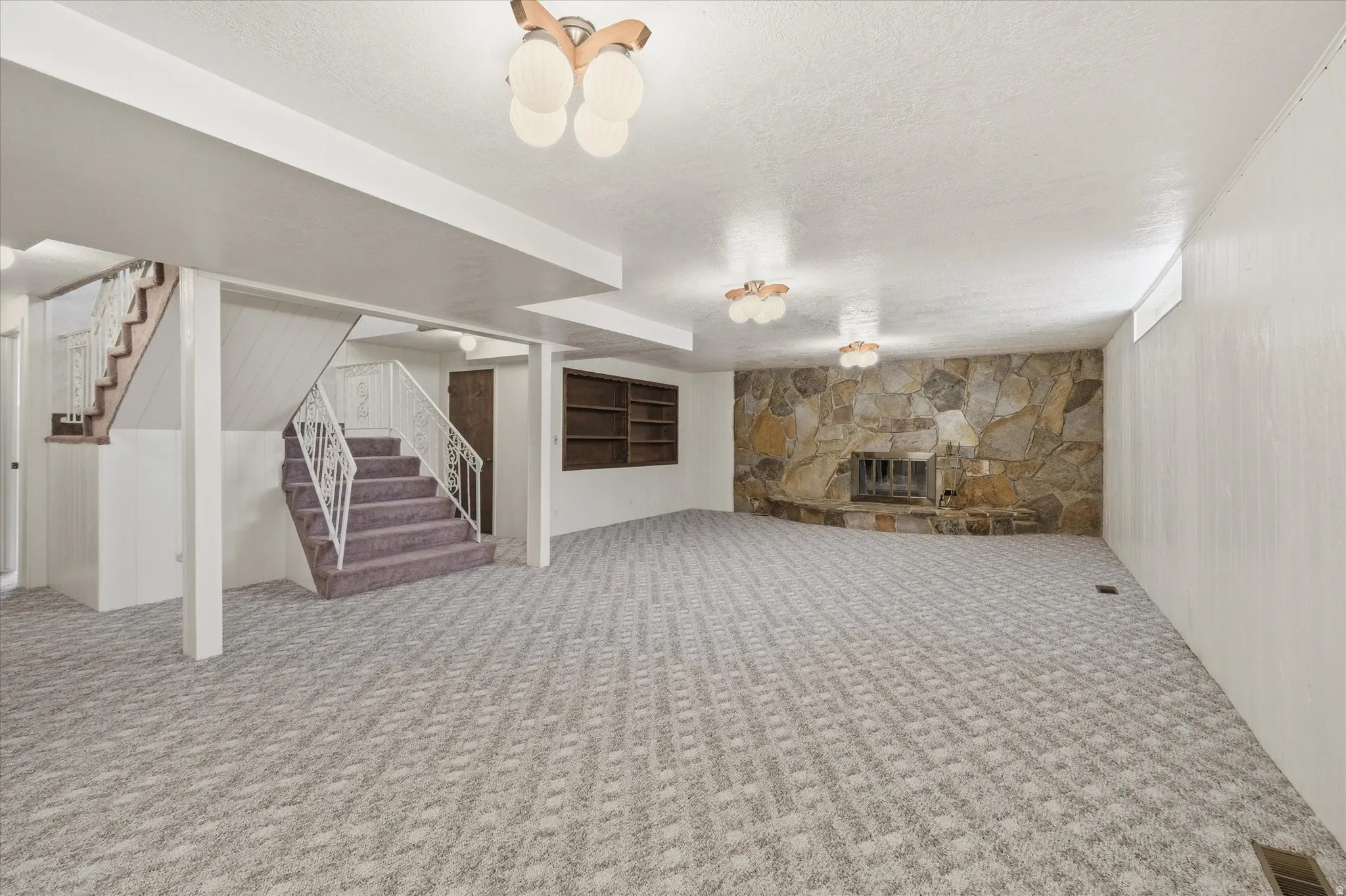 Spacious carpeted room featuring a stone-clad fireplace, integrated shelving, white paneled walls, and overhead lighting