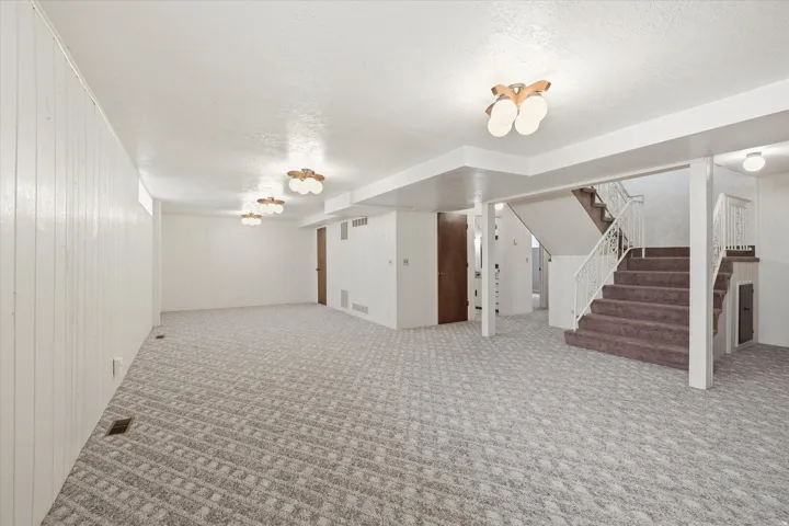 Spacious lower-level area with patterned carpet flooring, a carpeted staircase with a white metal railing, white paneled walls, and multiple ceiling-mounted light fixtures