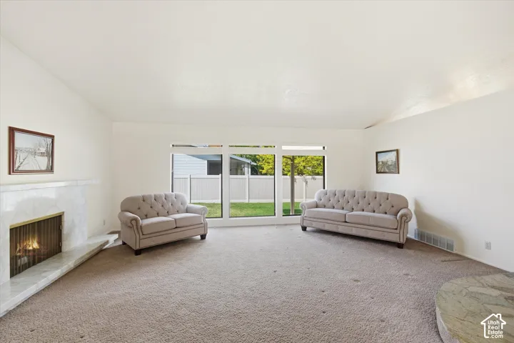 Spacious living area featuring a prominent fireplace with a marble-finish surround, large windows providing natural light, and plush carpet flooring throughout
