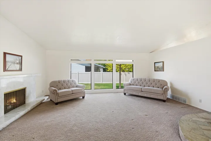 Spacious living area featuring a prominent fireplace with a marble-finish surround, large windows providing natural light, and plush carpet flooring throughout