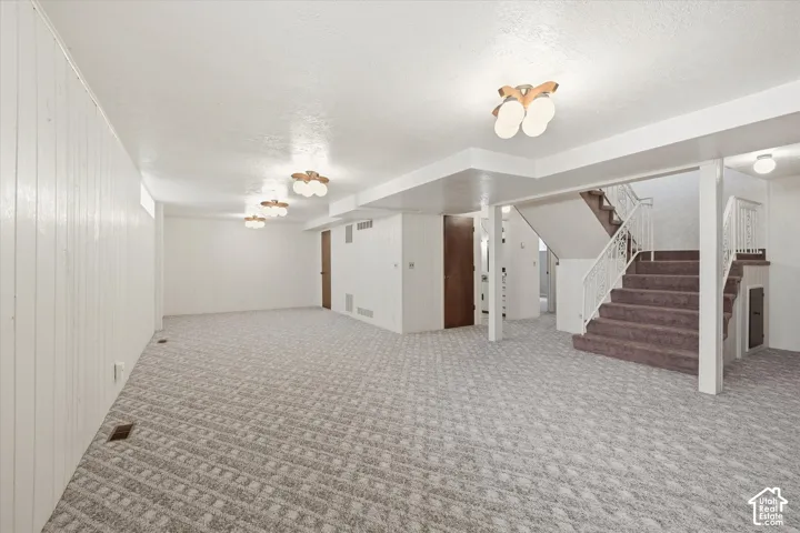 Spacious lower-level area with patterned carpet flooring, a carpeted staircase with a white metal railing, white paneled walls, and multiple ceiling-mounted light fixtures
