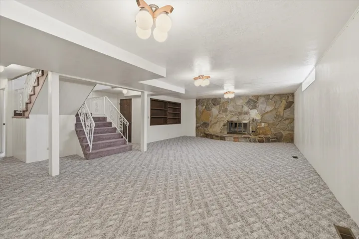 Spacious carpeted room featuring a stone-clad fireplace, integrated shelving, white paneled walls, and overhead lighting