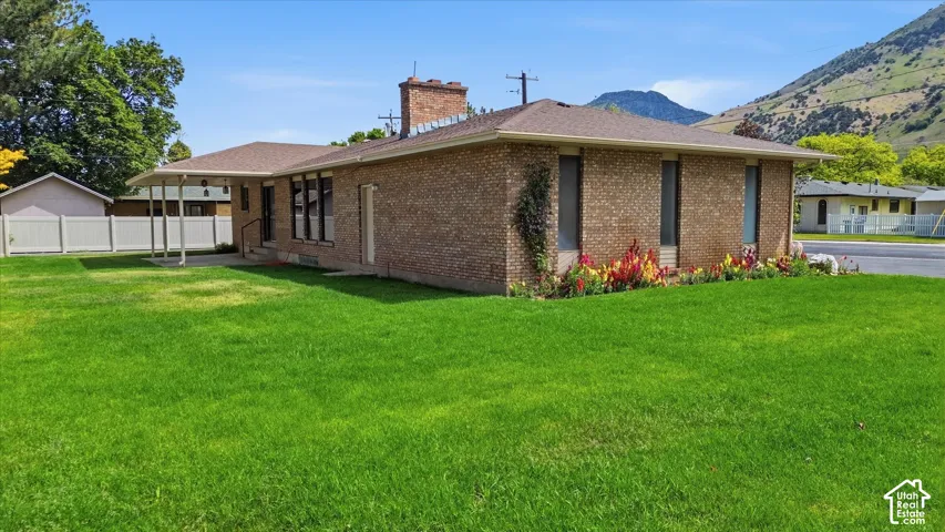 Brick exterior featuring a covered patio, extensive lawn space, mature trees, a garden bed with blooming plants, and a partial mountain backdrop