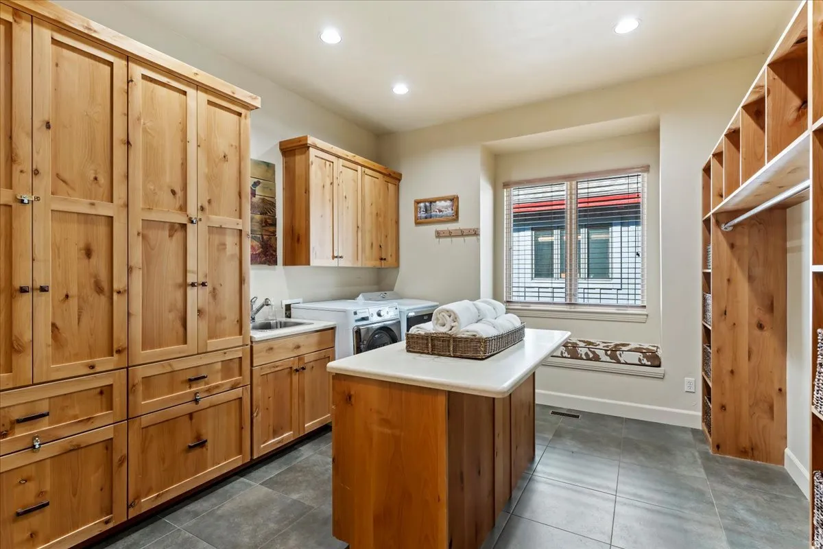 Spacious utility room featuring extensive natural wood cabinetry, a built-in sink, durable tile flooring, a functional center island with a light-colored countertop, and a window with a built-in bench