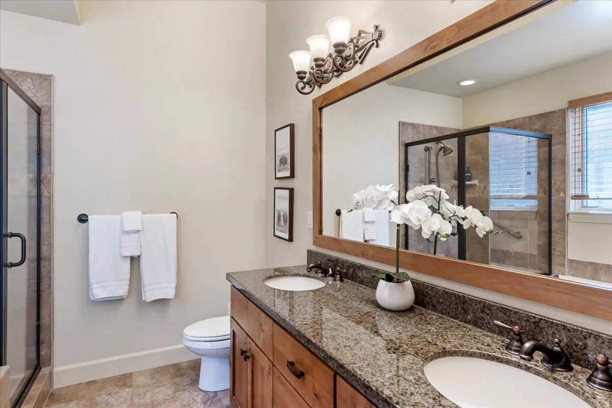 Spacious bathroom featuring a dual vanity with granite-style countertop, wood-finish cabinetry, a large framed mirror, and a glass-enclosed shower with tiled walls