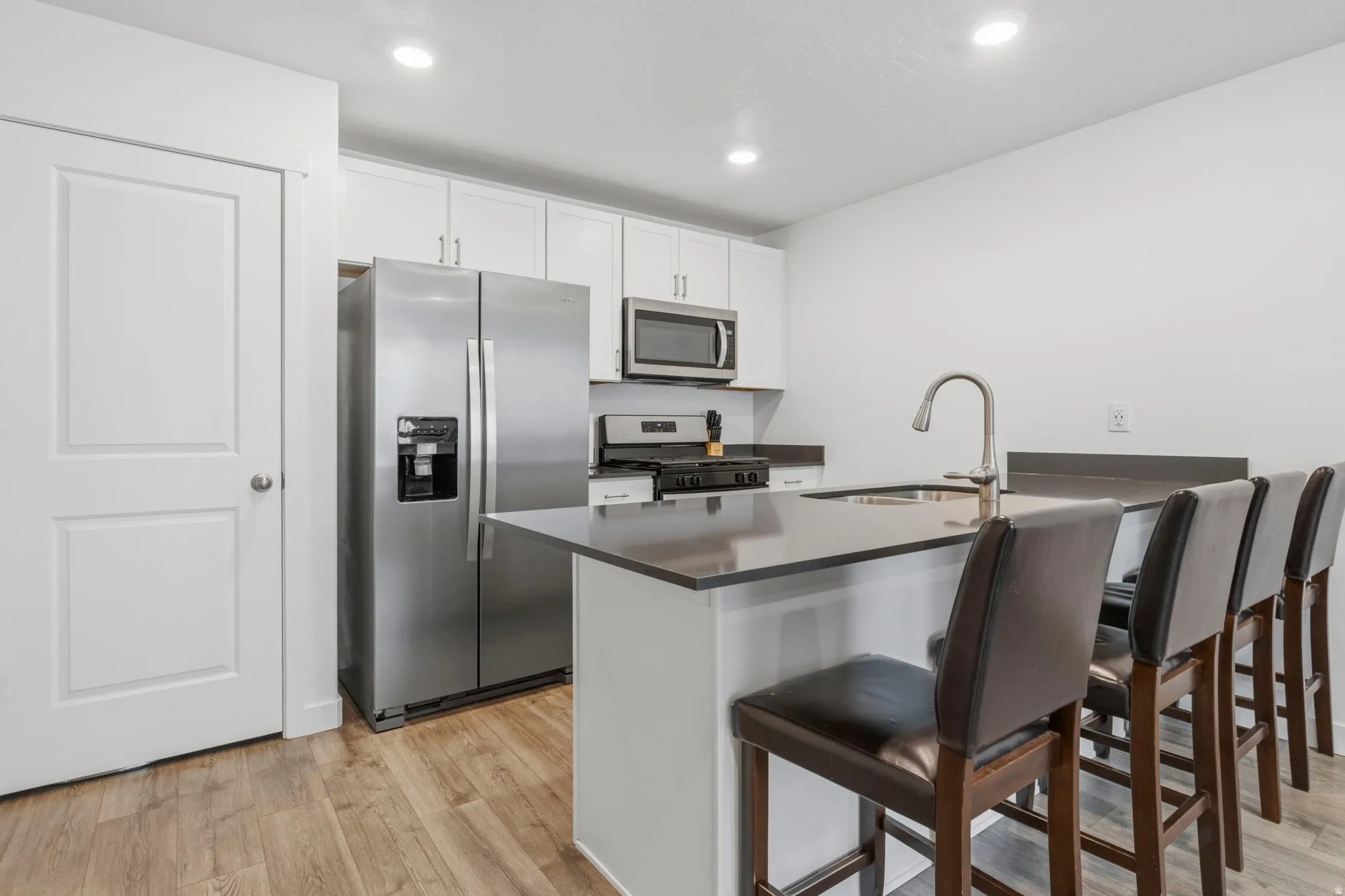 Modern kitchen featuring light wood-finish flooring, white cabinetry, stainless steel appliances, and a dark countertop island with an integrated sink