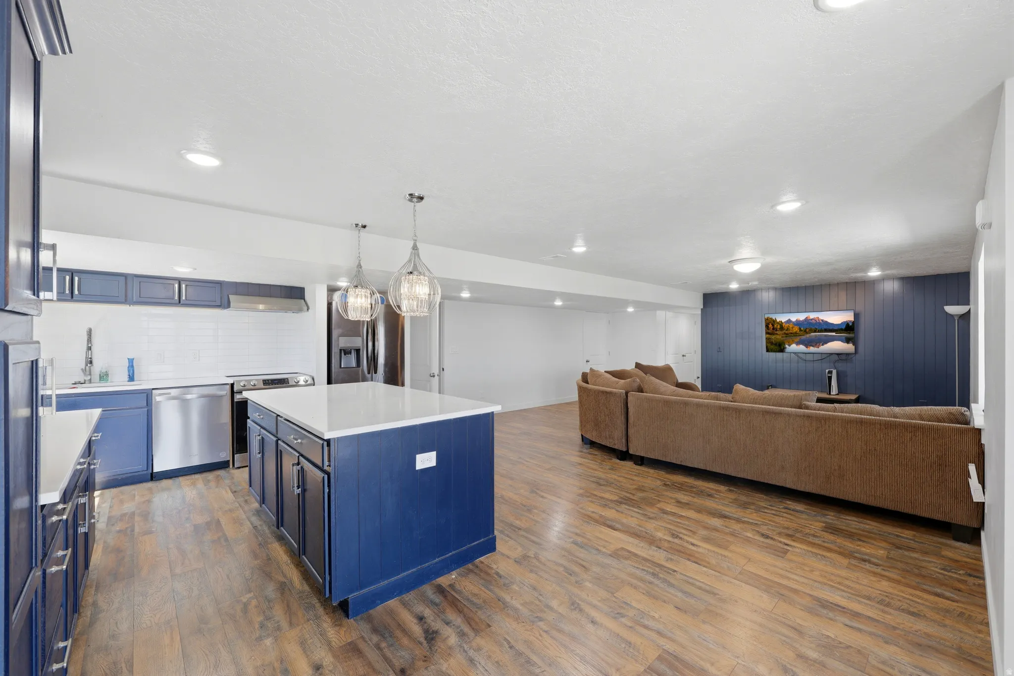 Open-concept living space with wood-finish flooring, a kitchen featuring blue cabinetry, white countertops, and stainless steel appliances, and recessed lighting throughout