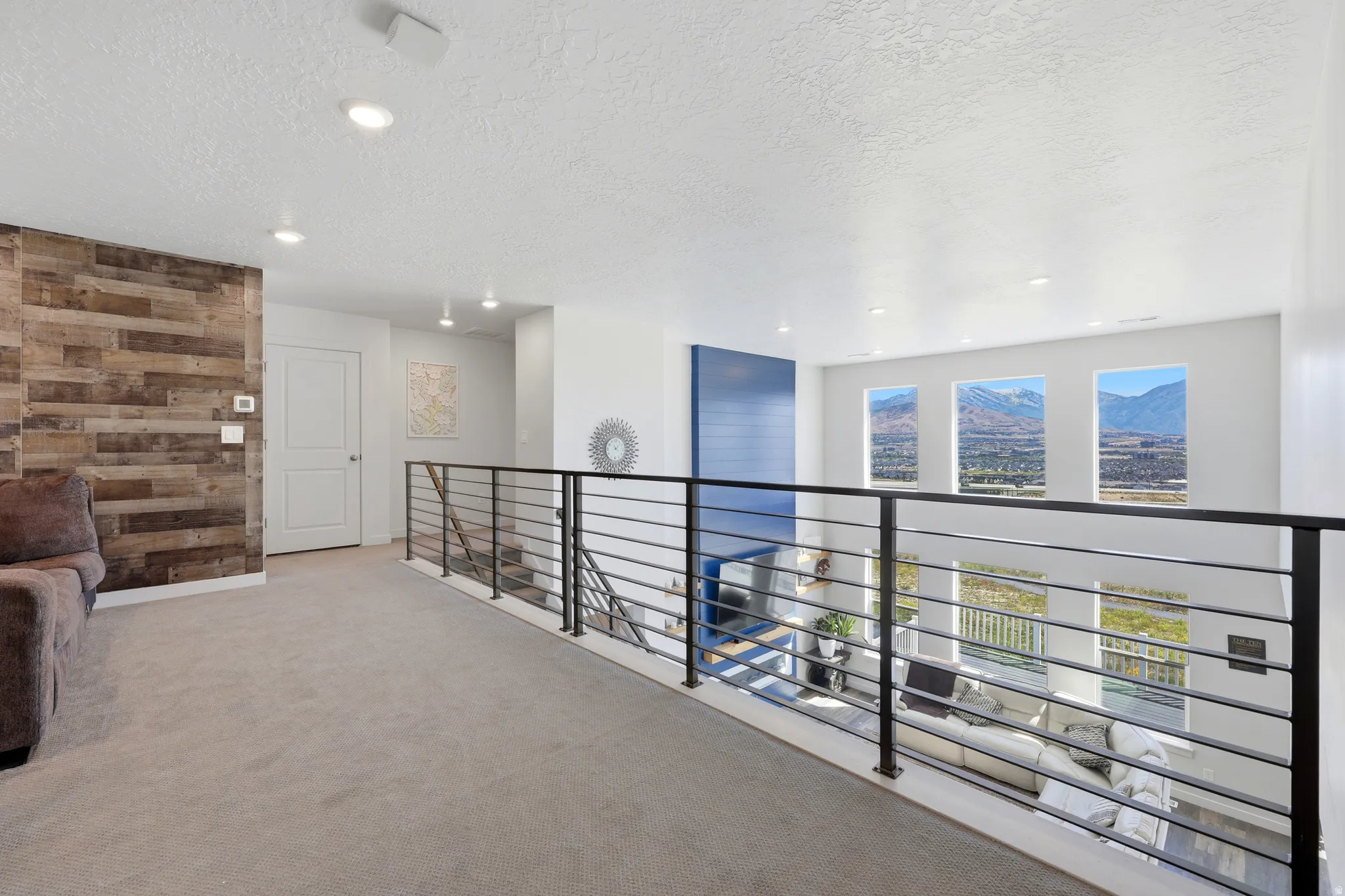 Carpeted loft space featuring a wood-finish accent wall, contemporary horizontal bar railing, recessed lighting, and three prominent windows showcasing distant mountains