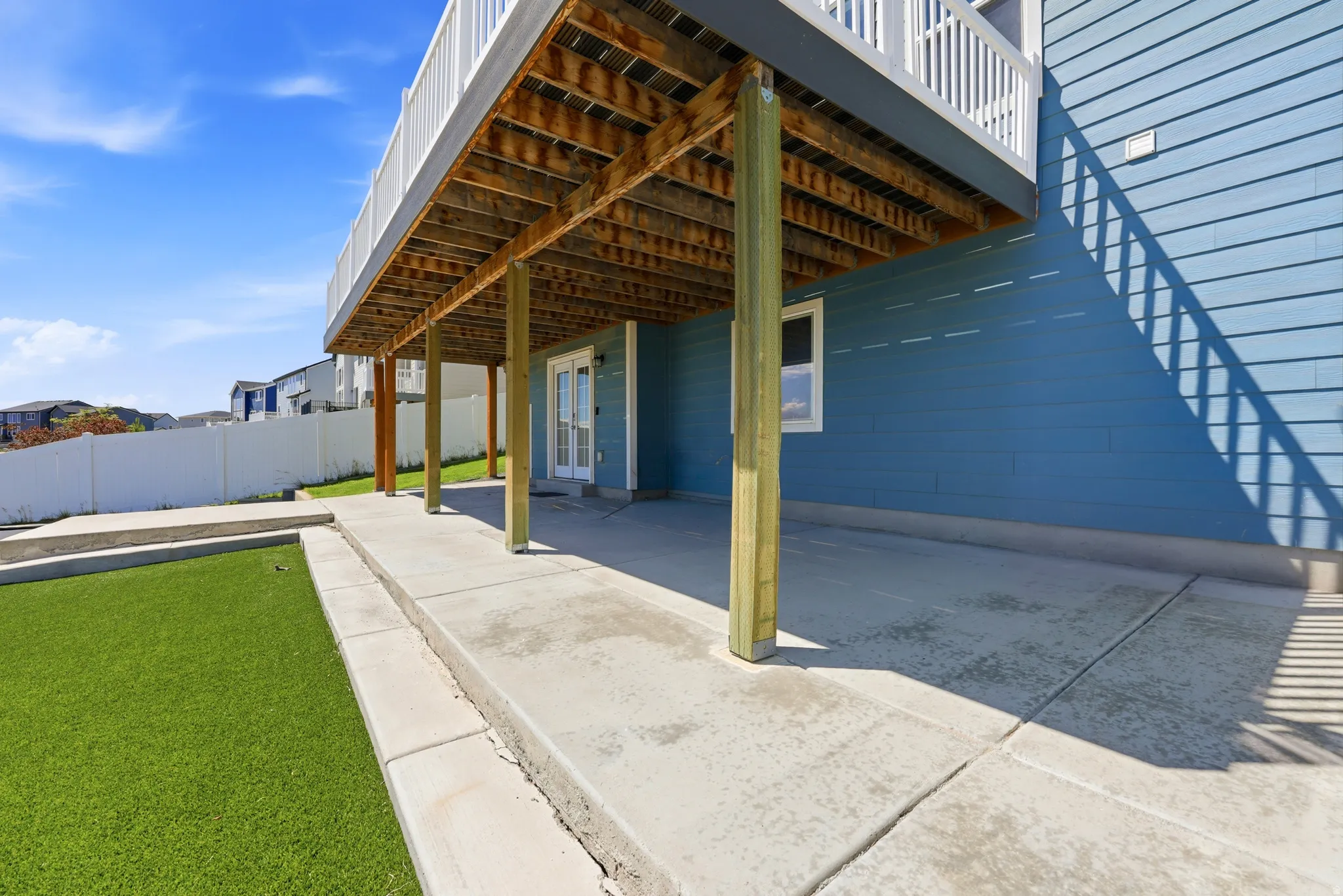 Lower patio area featuring concrete paving, an artificial turf section, and a deck overhead supported by wood posts