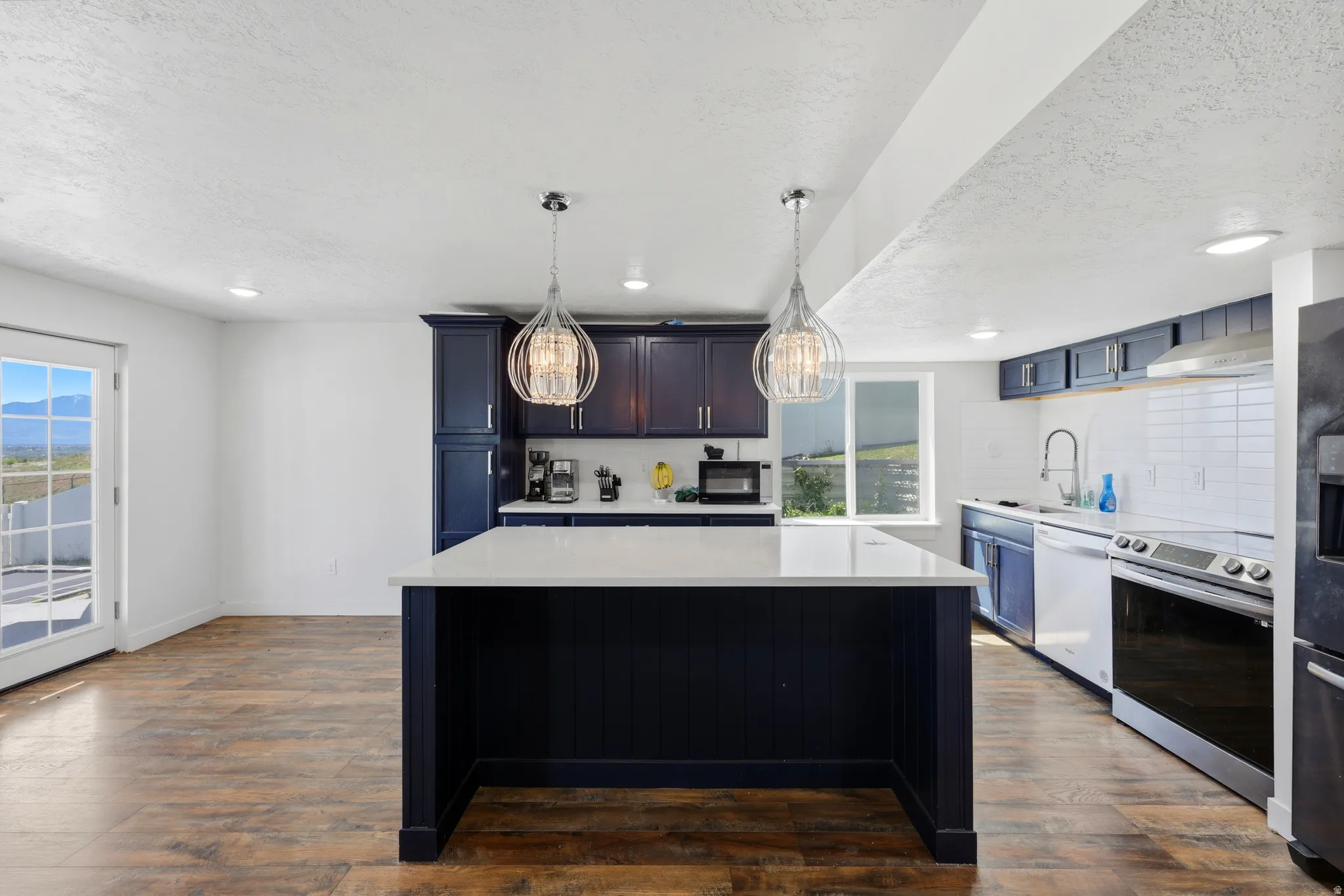 Open-concept kitchen featuring a dark blue island with a light-toned countertop, two decorative pendant lights, stainless steel appliances, and wood-finish flooring