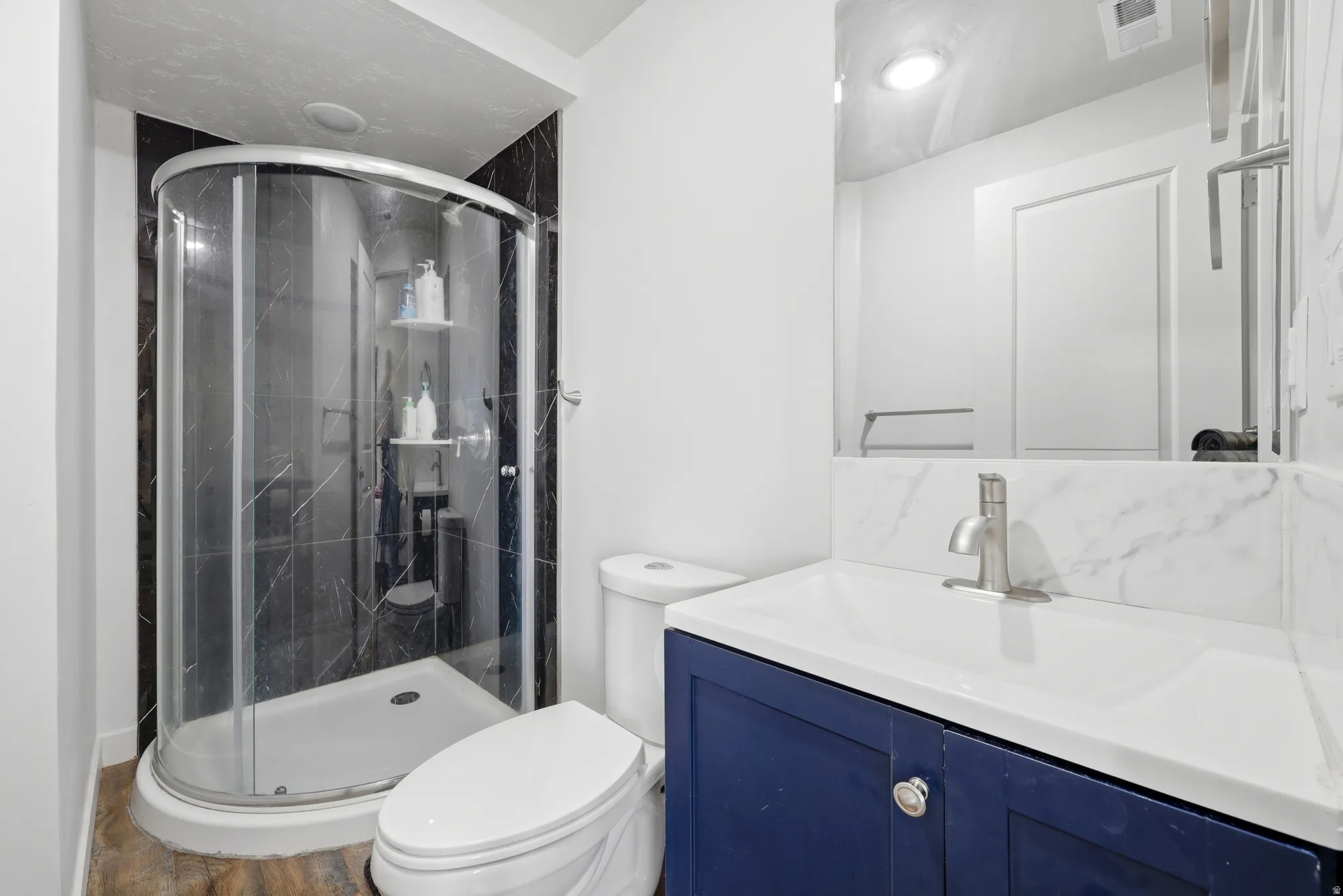 Three-piece bathroom featuring a corner shower with dark tile surround, a single-sink vanity with blue cabinetry, and a white countertop with a matching backsplash