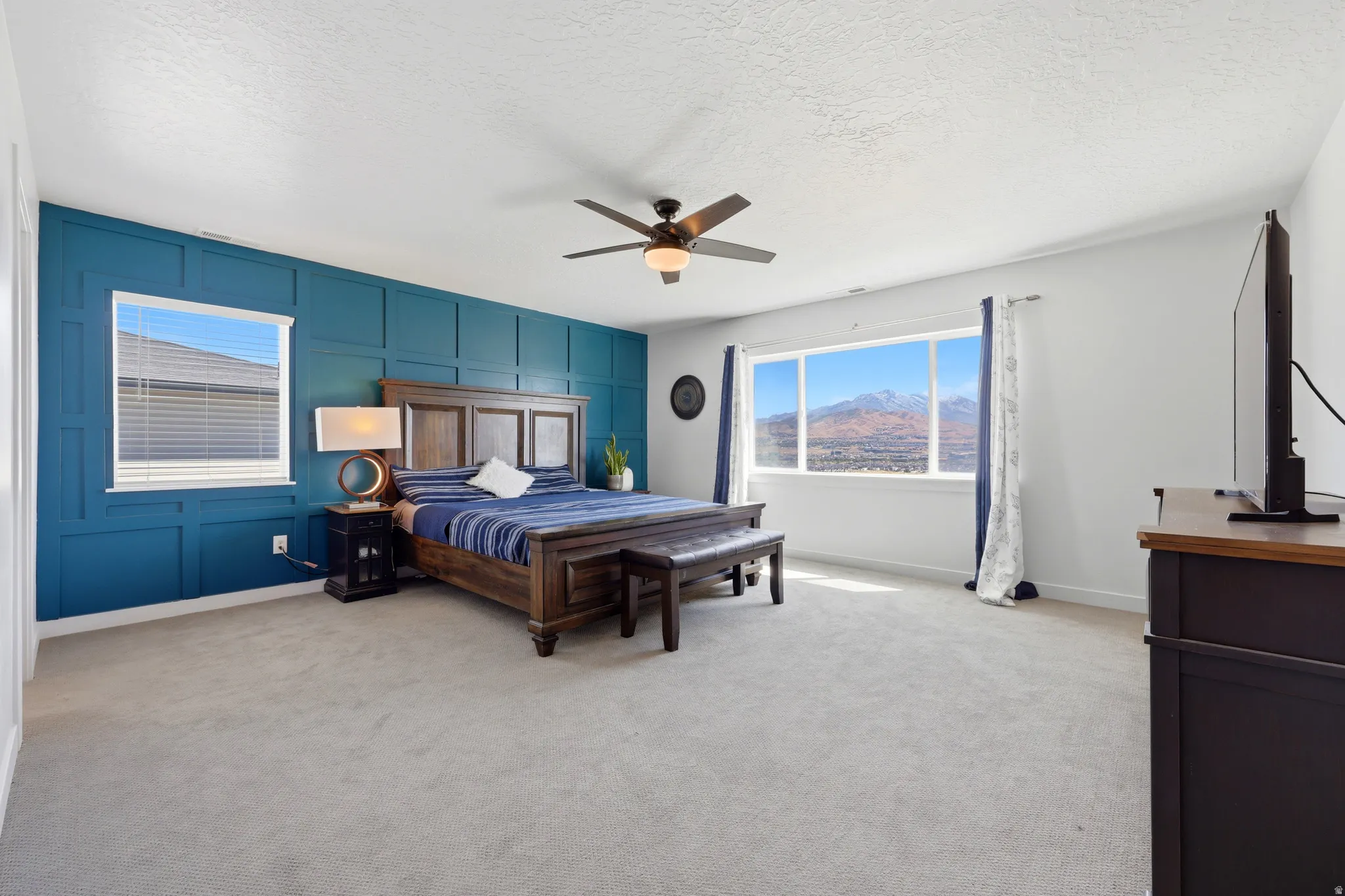 Spacious bedroom featuring a paneled accent wall, a wide window with mountain scenery, a ceiling fan, light gray carpeting, and white walls