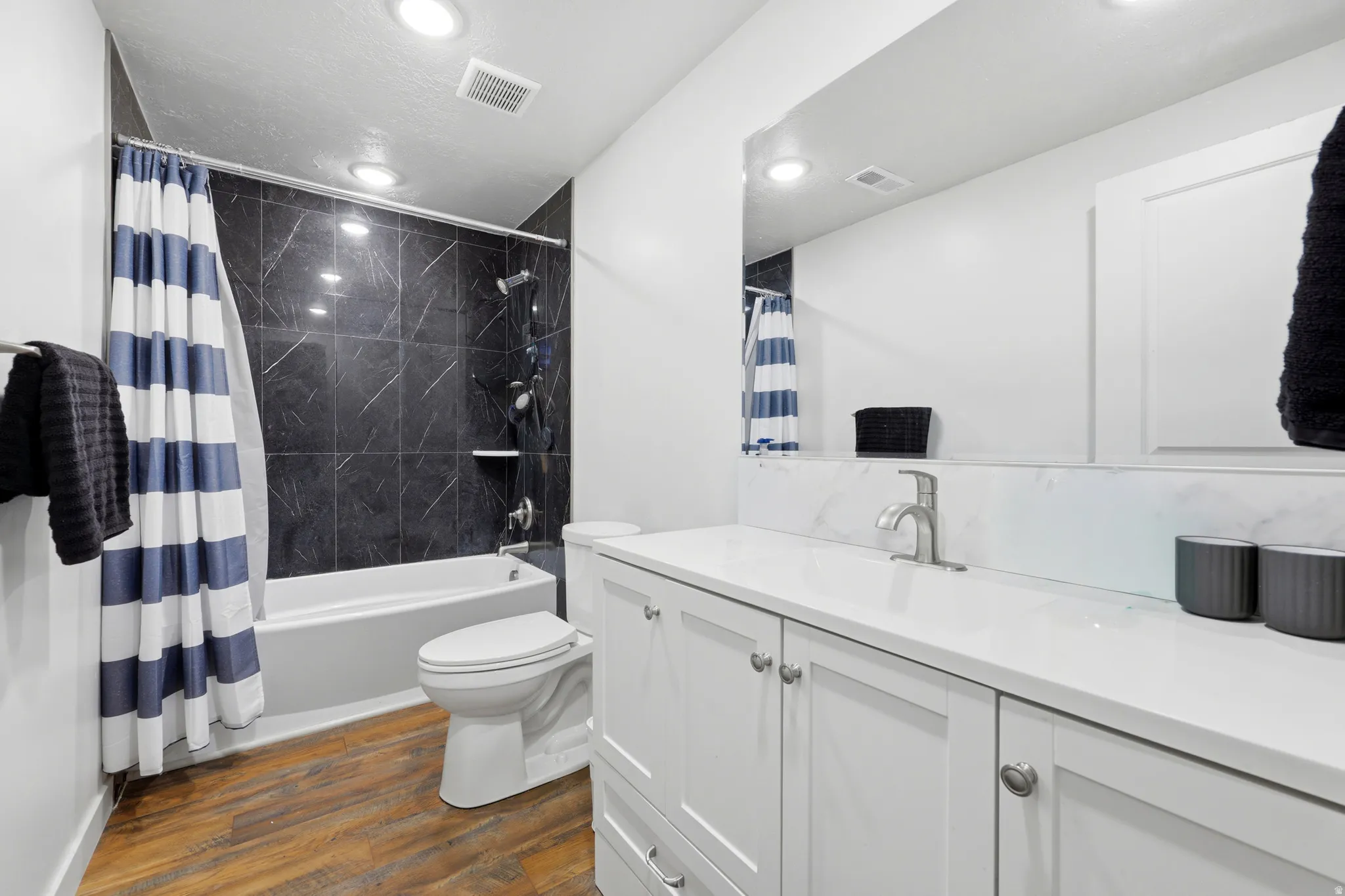 Bathroom featuring a white vanity with a light-colored countertop and integrated sink, a large mirror, a toilet, and a bathtub with dark stone-look tile surround