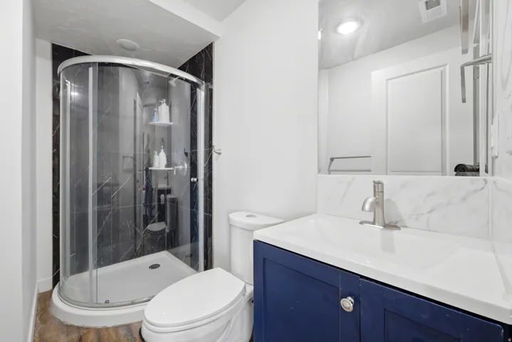 Three-piece bathroom featuring a corner shower with dark tile surround, a single-sink vanity with blue cabinetry, and a white countertop with a matching backsplash