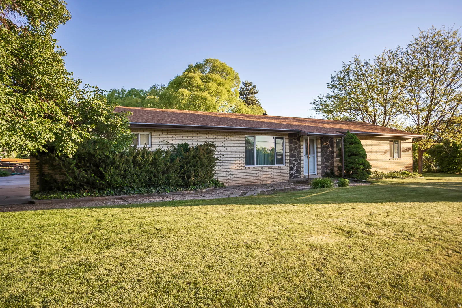 Front exterior featuring light brick and stone veneer, a brown shingled roof, a paved walkway, and mature landscaping