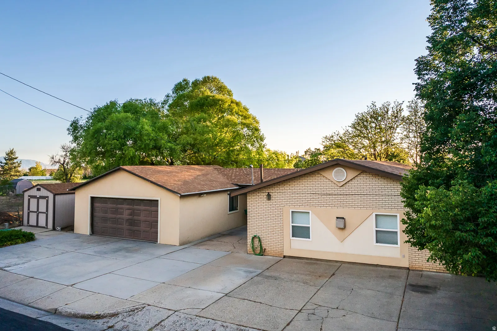 Exterior featuring a tan stucco finish with light brick accents, a brown shingle roof, a two-car garage, and an additional detached shed