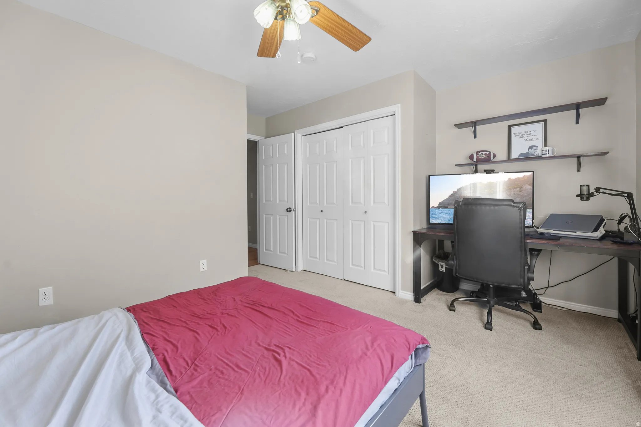 Carpeted room with a ceiling fan featuring wood-finish blades, a double-door closet with louvered panels, and a built-in wall shelf system