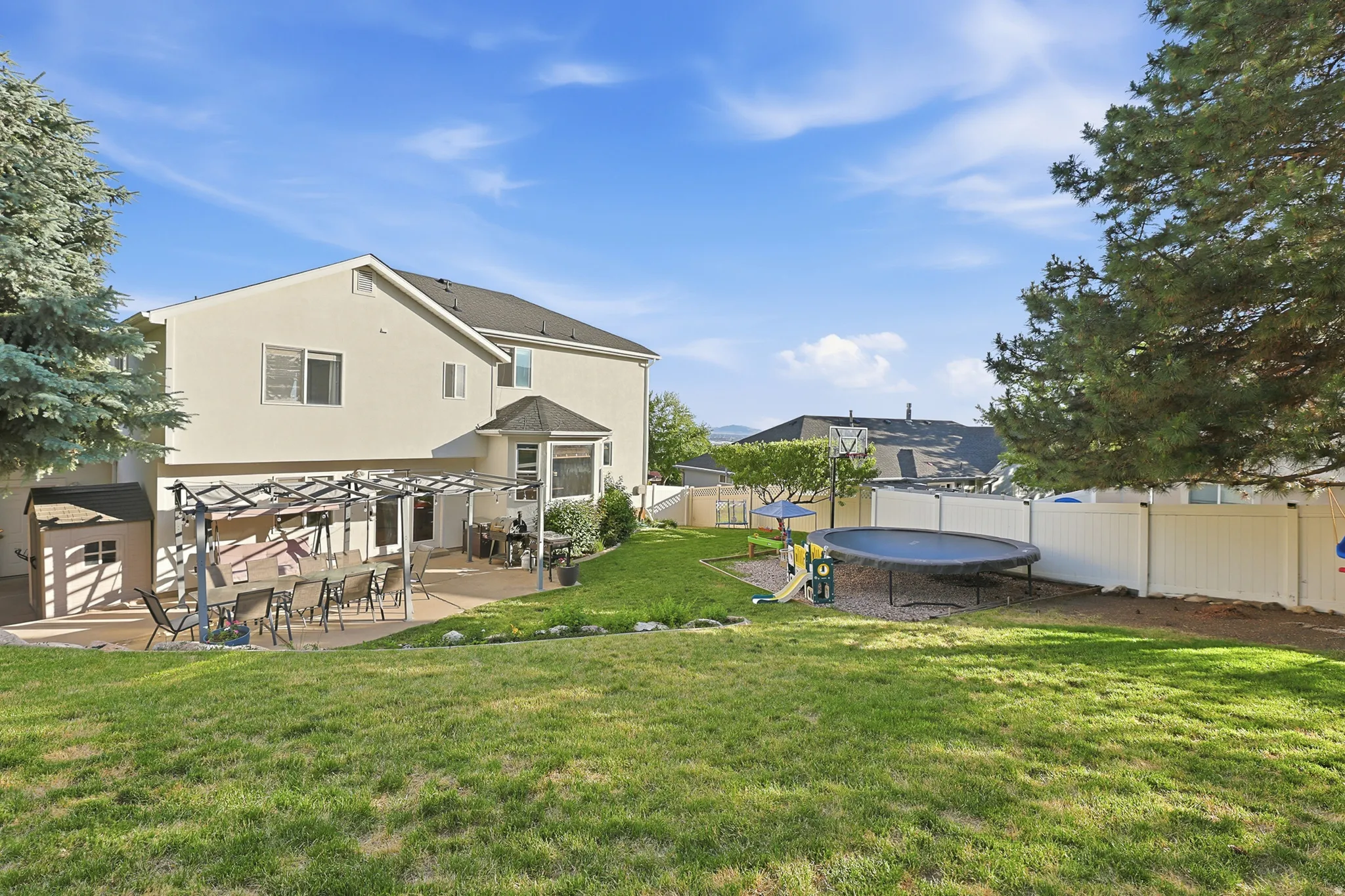 Expansive backyard featuring a large concrete patio with a pergola, a spacious lawn, a white privacy fence, and mature evergreen trees