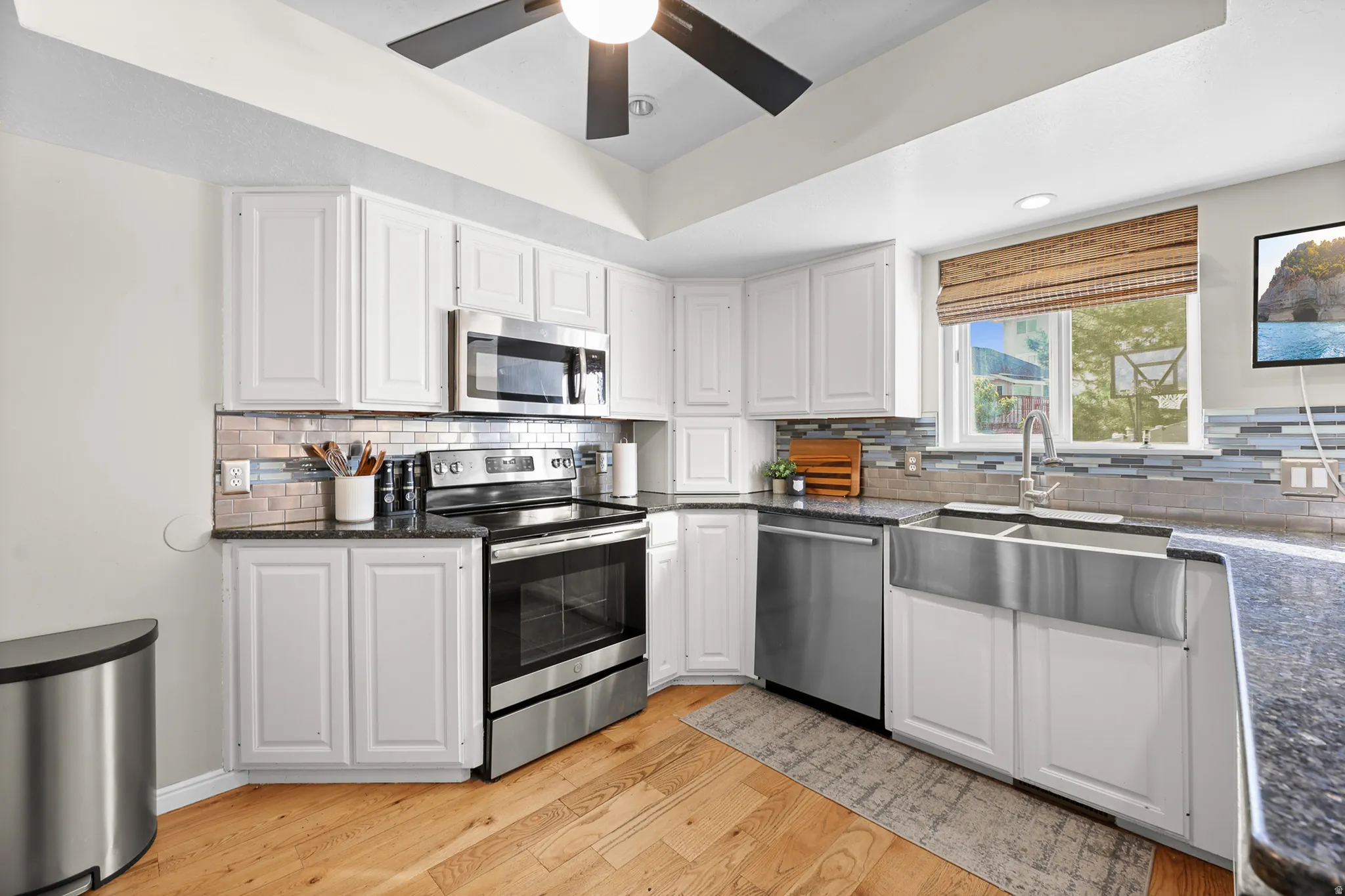 Modern kitchen featuring white cabinetry, dark speckled countertops, stainless steel appliances, a farmhouse-style sink, and wood-finish flooring