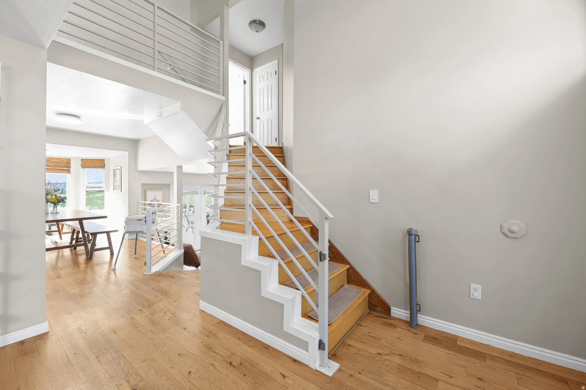Open-concept interior featuring light wood-finish flooring, a contemporary staircase with horizontal metal railings, and a bay window in the background