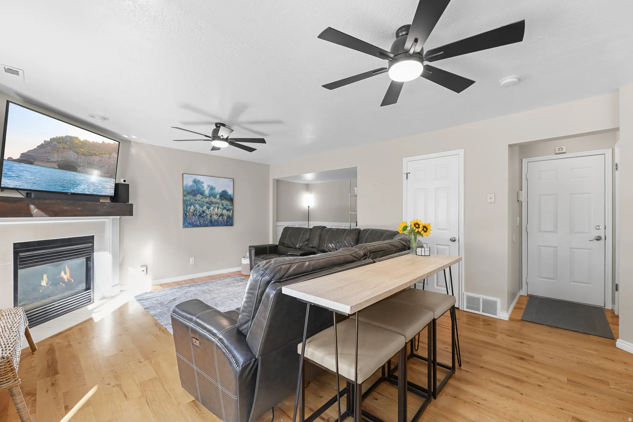 Open-concept living area with wood-finish flooring, a white mantel fireplace, and two ceiling fans with integrated lighting