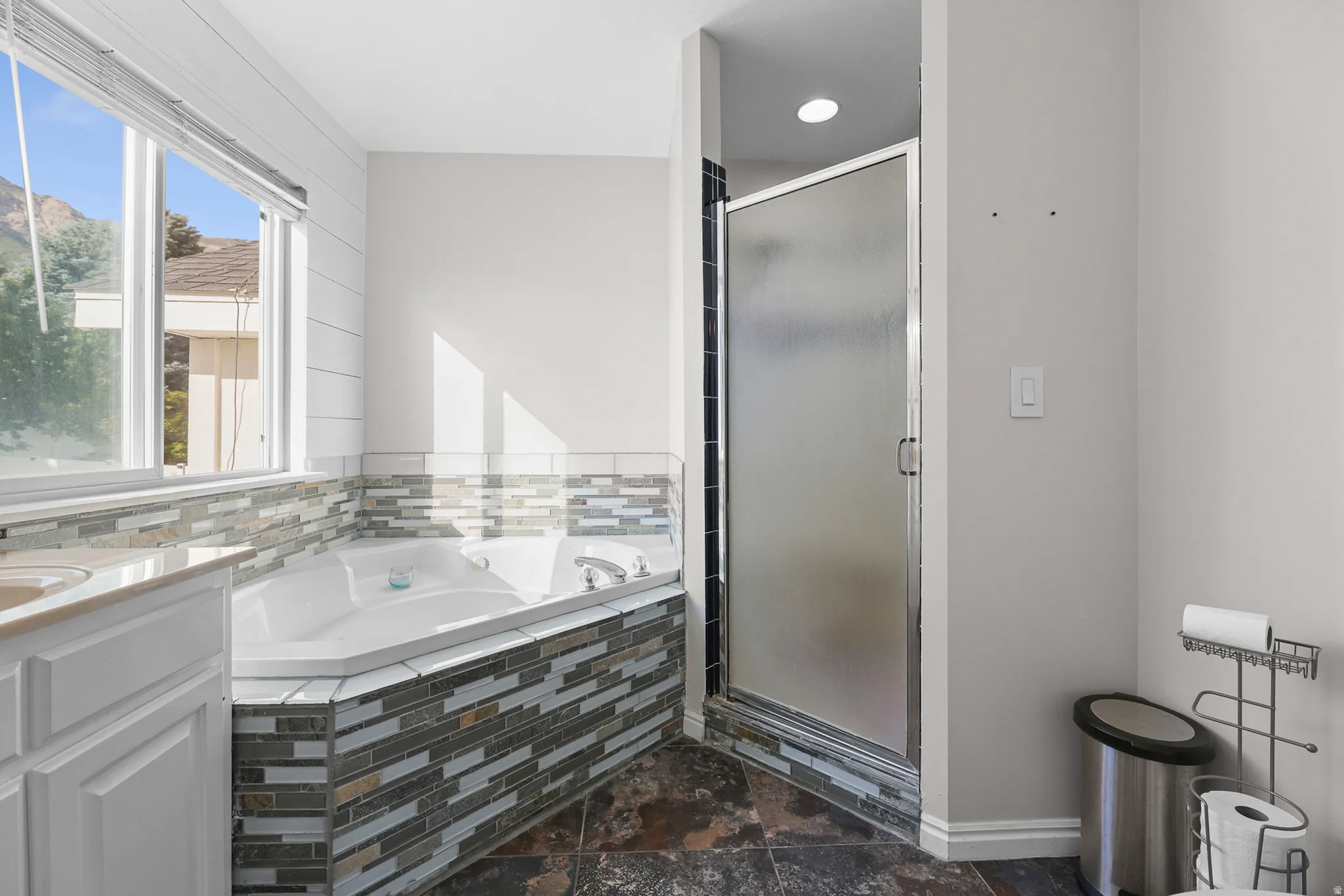 Bathroom featuring a corner jetted tub with tile surround, a separate glass-enclosed shower, a vanity with a countertop, and large format floor tiling