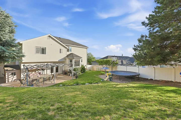 Expansive backyard featuring a large concrete patio with a pergola, a spacious lawn, a white privacy fence, and mature evergreen trees