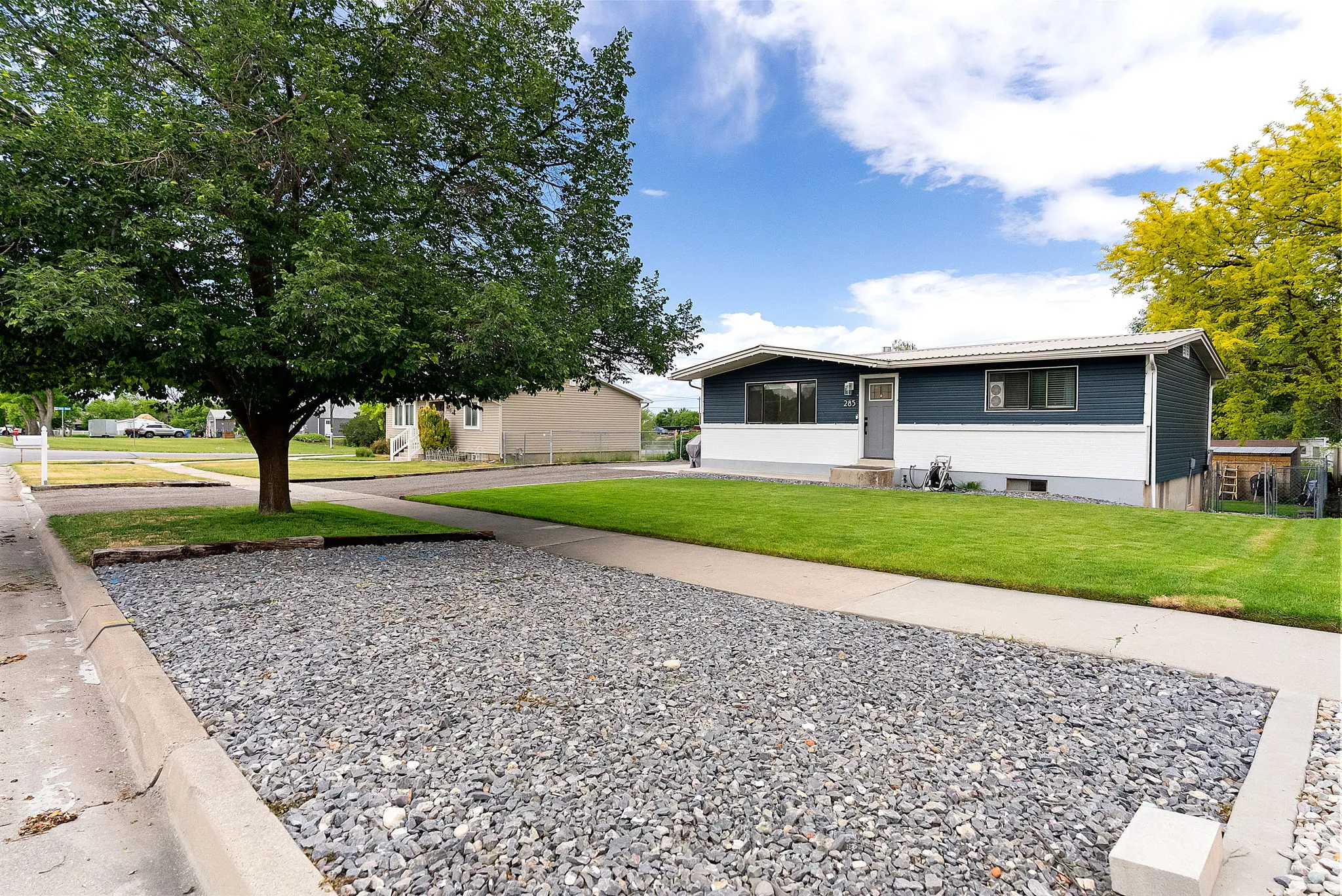 Split-level residence featuring a blue and white exterior, a front yard with green lawn, and a tree-shaded gravel parking area