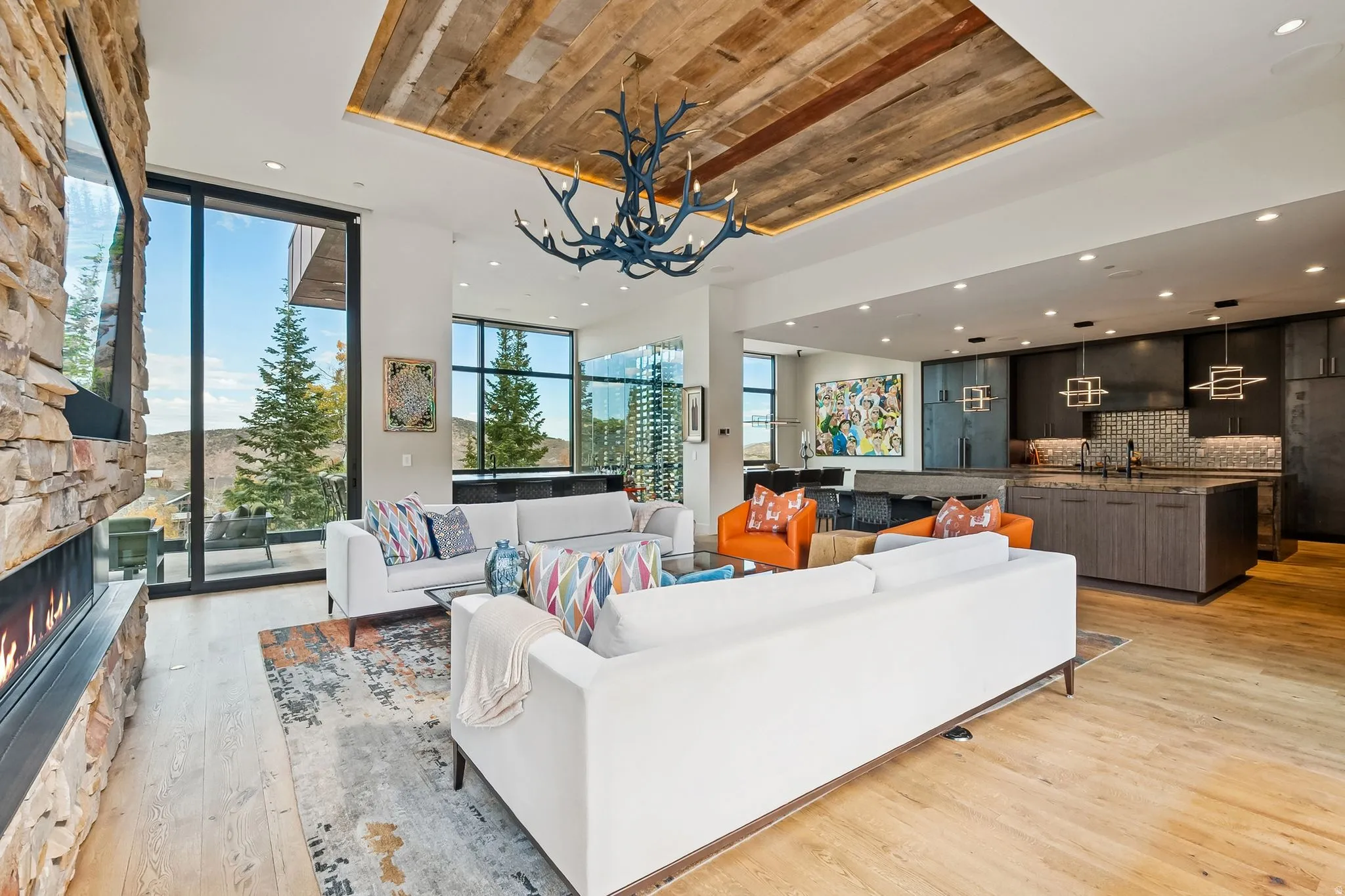 Living area featuring light wood-style flooring, a stone fireplace, expansive windows, suspended lighting, and a wood tray ceiling