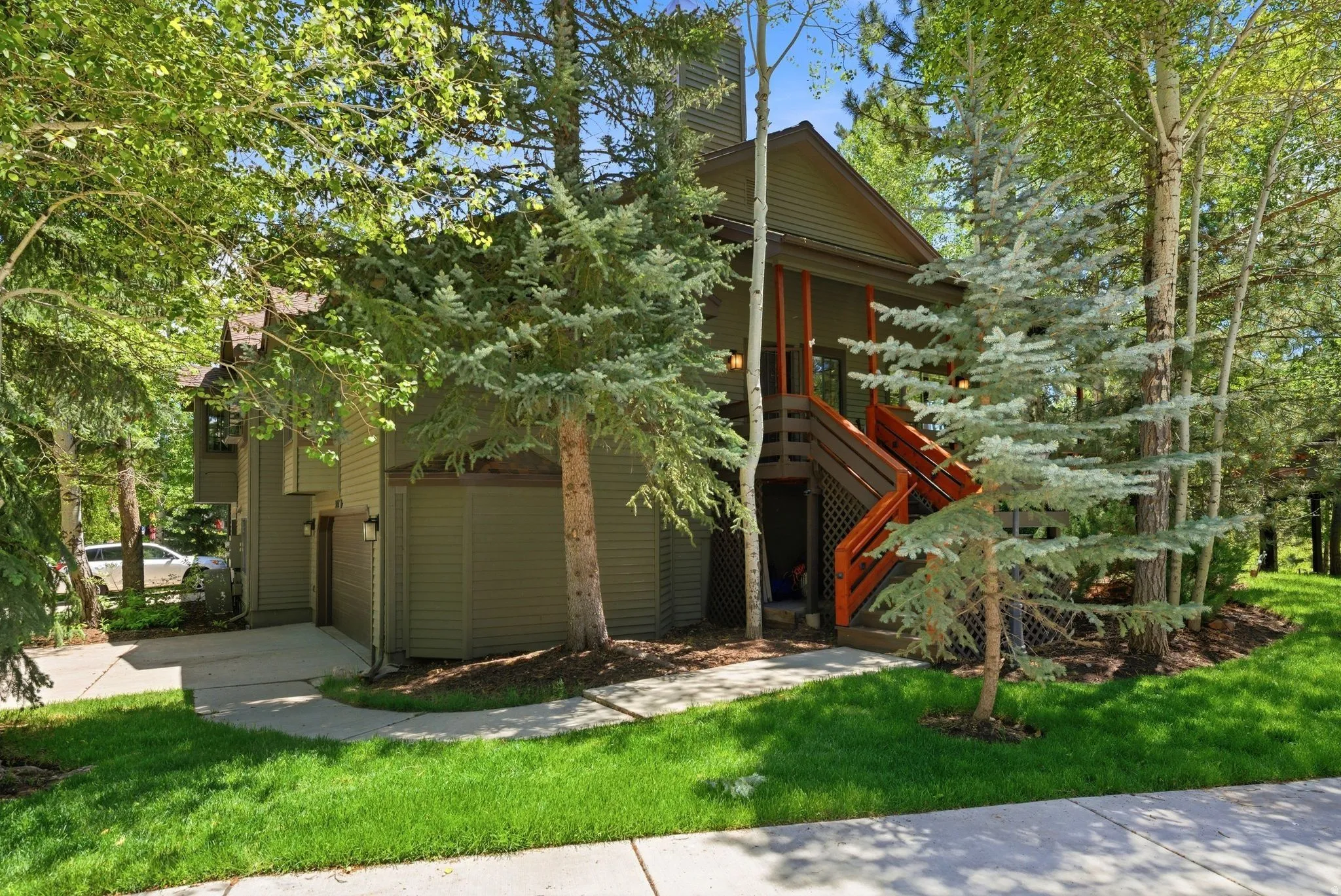 Multi-level exterior with olive green siding, a prominent staircase with orange-stained railings, a pitched roof, and lush surrounding landscaping including mature deciduous and evergreen trees