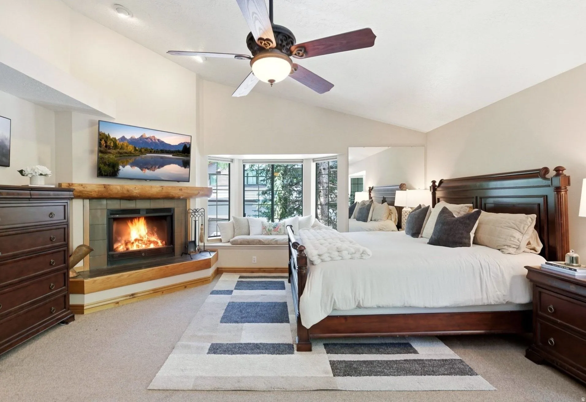 Vaulted ceiling room featuring a corner fireplace with a wood mantle and tile surround, a built-in window seat, a ceiling fan with integrated lighting, and neutral-toned carpeting