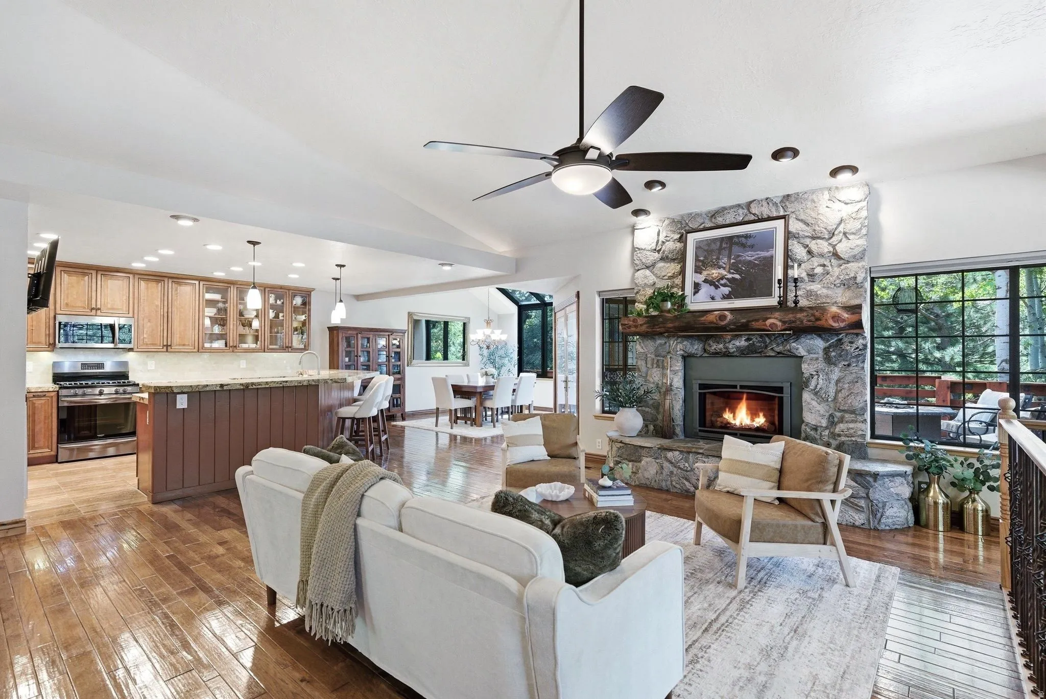Open-concept living area featuring wood-finish flooring, a stone-surround fireplace with a substantial timber mantel, and large picture windows