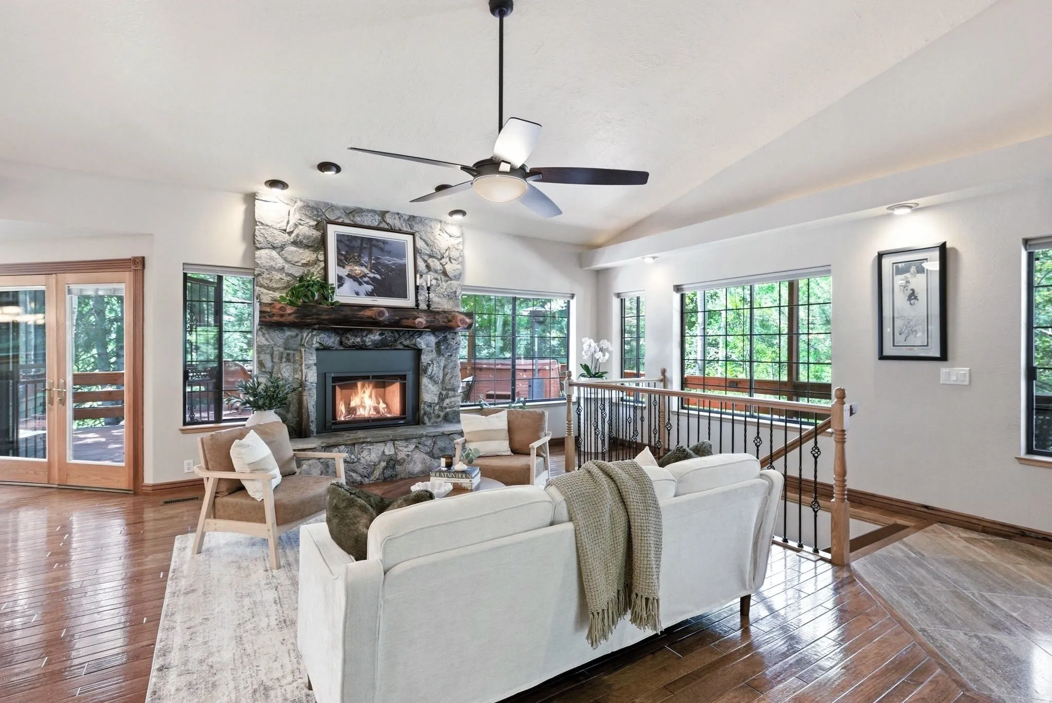 Spacious living area featuring a stone-faced fireplace with a substantial timber mantel, wood-finish flooring, a vaulted ceiling, and multiple windows